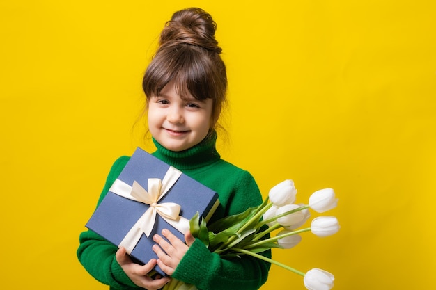 Uma menina morena feliz está segurando uma caixa de presentes e um buquê de tulipas em um fundo amarelo em