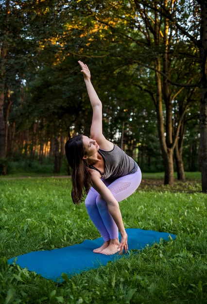 Uma menina morena fica em uma pose de ioga em um tapete azul na floresta ao pôr do sol