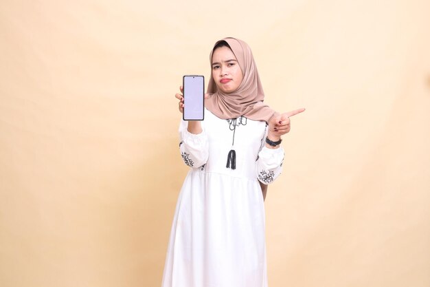 Uma menina muçulmana indonésia vestindo um hijab franzeia a testa mostrando um aparelho de tela de celular apontando para a esquerda