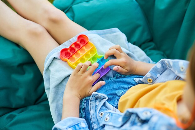 Uma menina na cadeira de suspensão ao ar livre jogar pop, mãos de criança jogando bolhas de um brinquedo de destressão do arco-íris, brinquedo de agitação no quintal da casa em um dia ensolarado de verão, férias de verão.