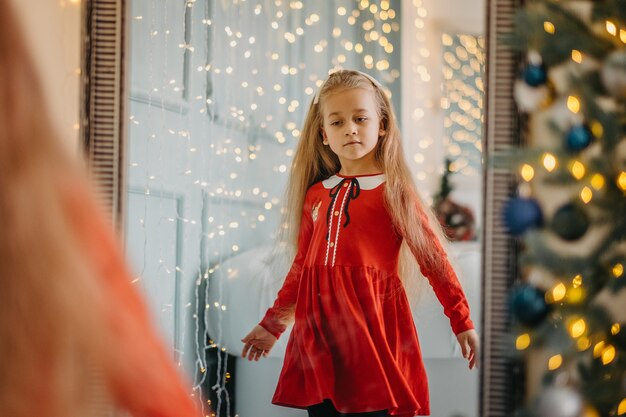 Uma menina na véspera de Natal com roupas festivas fica perto do espelho e olha para seu reflexo. A criança se olha no espelho antes do ano novo.