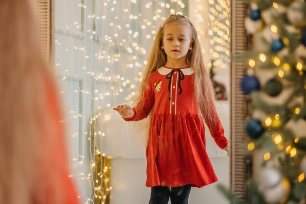 Uma menina na véspera de Natal com roupas festivas fica perto do espelho e olha para seu reflexo. A criança se olha no espelho antes do ano novo.