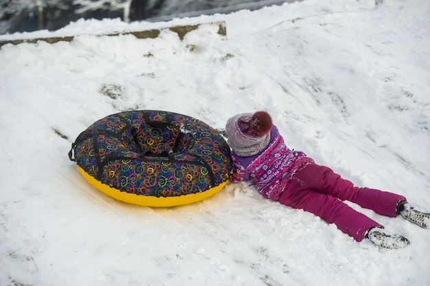Uma menina no inverno com roupas roxas e um círculo inflável desce a colina na rua