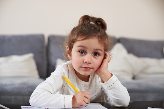 Uma menina olha para a câmera, apoiando a cabeça na palma da mão. Garota desenhando em casa