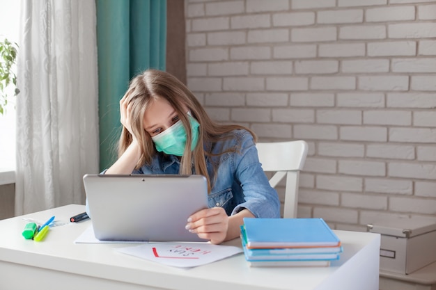 Uma menina pensativa em uma máscara médica está estudando em casa com um laptop tablet digital e fazendo lição de casa. ensino a distância, educação on-line