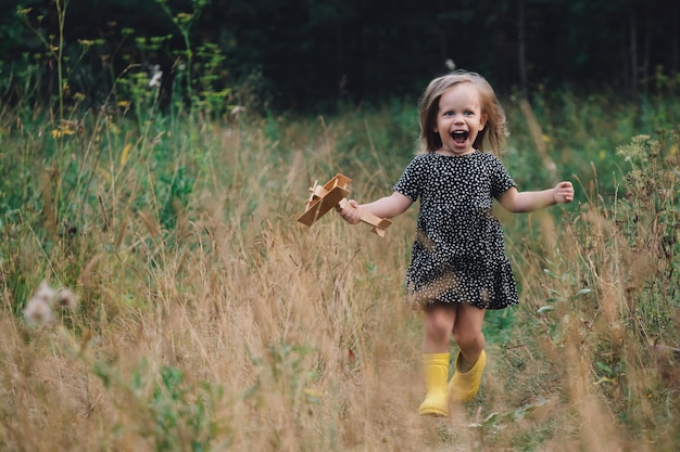 Uma menina pequena está brincando com um avião de madeira na natureza liberdade sonho imaginação conceito infância feliz uma menina bonita corre alegremente ao longo de um caminho na grama ri sorrisos cópia espaço