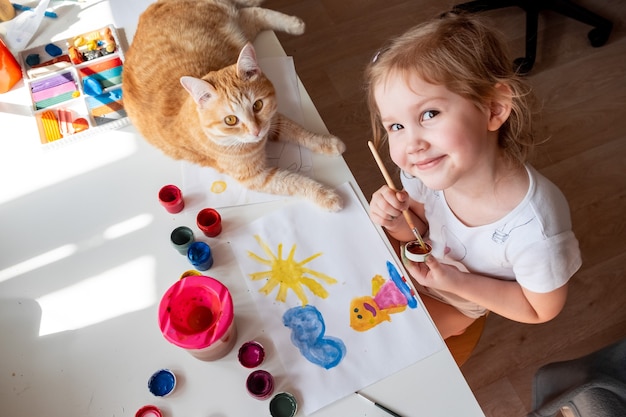 Uma menina pinta o sol e sua mãe com aquarelas um gato ruivo deita ao lado da mesa.