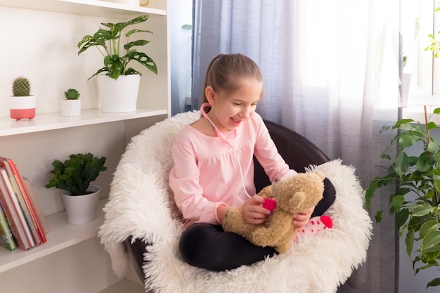 Uma menina pré-escolar vestida com uma camiseta rosa toca em casa em uma poltrona no hospital ouve um