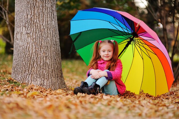 Uma menina ruivo pequena em um revestimento cor-de-rosa senta-se nas folhas amarelas com um grande guarda-chuva da cor do arco-íris.