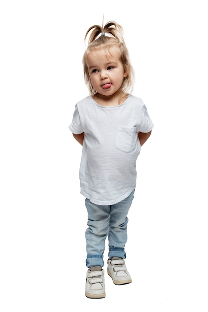 Uma menina se levanta e mostra a língua. Criança de 4 anos vestindo jeans e uma camiseta branca. Altura toda. Isolado em um fundo branco. Vertical.
