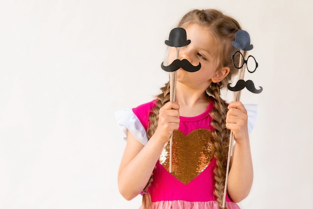Uma menina segura um bigode de carnaval, chapéu e óculos para o dia dos pais.