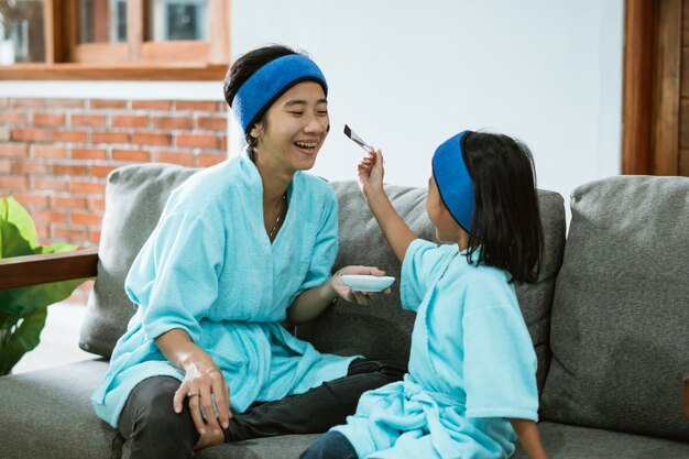 Uma menina segurando uma escova facial esfregou o rosto da mãe com uma máscara de argila quando estava sentada no sofá