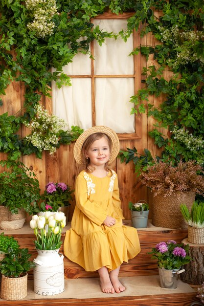 Uma menina senta-se na varanda da casa de madeira, em torno de plantas e flores verdes. garoto está vestido amarelo, chapéu de palha. conceito de infância. jardinagem. terraço rústico, alpendre. criança brincando no quintal