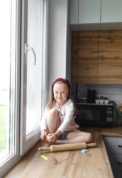 Uma menina senta-se no parapeito da janela da cozinha e brinca com os utensílios da cozinha