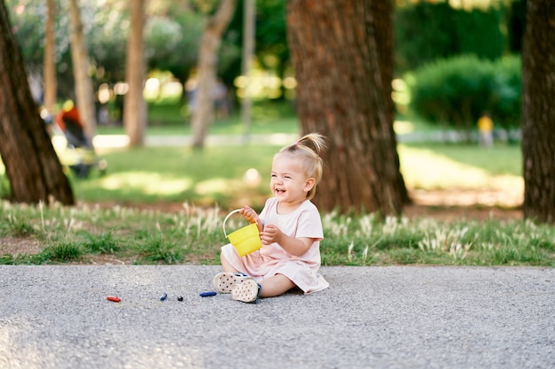 Foto uma menina sentada em um caminho de asfalto em um parque verde com um balde de brinquedo nas mãos