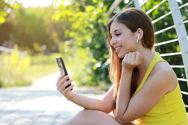 Foto uma menina sentada no parque a desfrutar de ouvir música usando um telemóvel com fones de ouvido sem fios