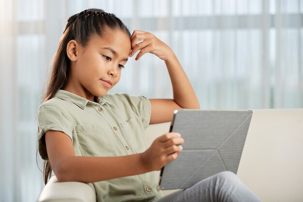 Uma menina sentada no sofá com um tablet digital, ela lendo um livro online durante seu tempo de lazer em casa