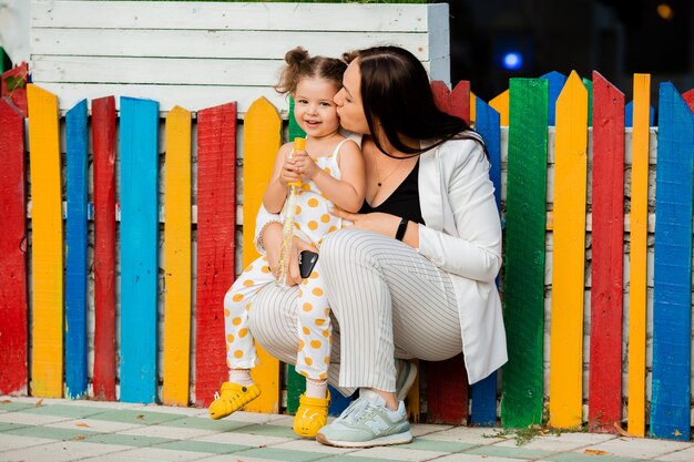 Uma menina sentada perto de uma cerca de madeira multicolorida com a mãe