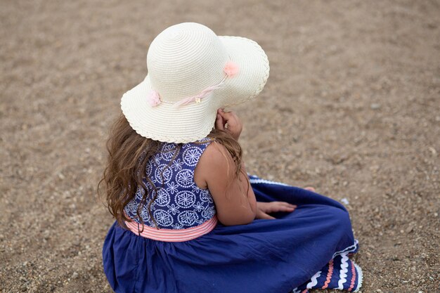 Uma menina sentada, segurando um chapéu de verão e um lindo vestido azul na praia