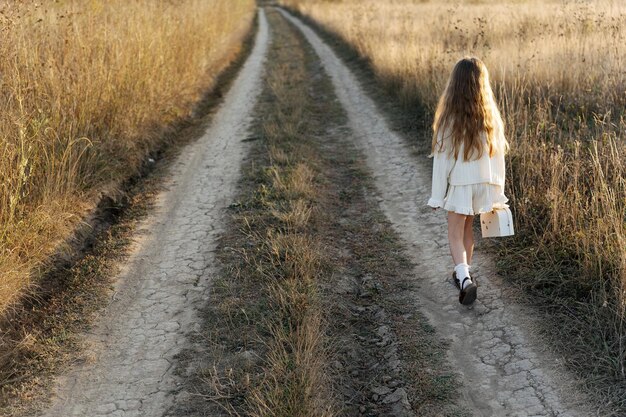 uma menina solitária em roupas leves com uma pequena mala de brinquedo no meio de uma estrada vazia em um campo