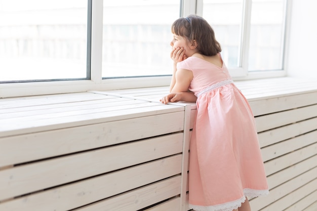 Uma menina sonhadora com um vestido rosa olhando pela janela