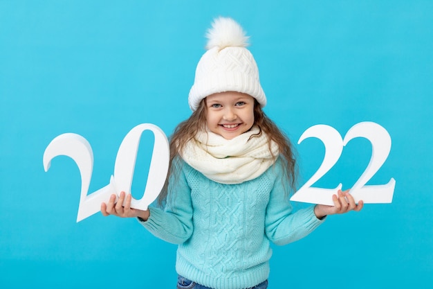 Uma menina sorridente com roupas de inverno, chapéu e suéter sobre um fundo azul isolado mostra os números do ano novo de 2022 e sorri, um lugar ou espaço para texto