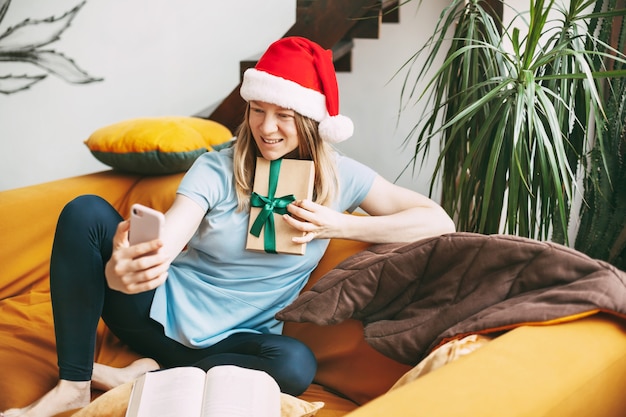 Uma menina sorridente com um chapéu de natal se senta no sofá e se comunica por meio de um link de vídeo com os amigos