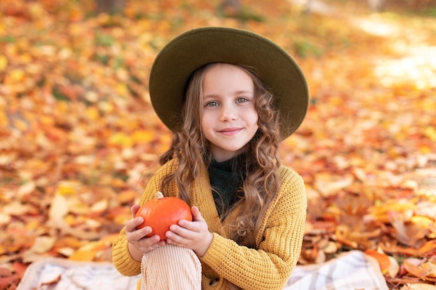 Foto uma menina sorridente de chapéu se diverte brincando com folhas douradas caindo e abóbora no outono