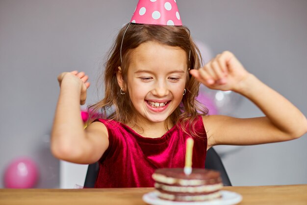 Uma menina sorridente e feliz está usando um chapéu de aniversário olhando para um bolo de aniversário, velas sopradas