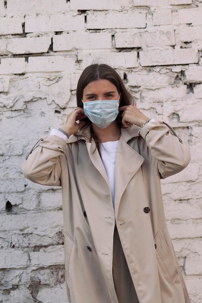 Foto uma menina vestida com roupas brancas quentes de pé contra uma parede de tijolos coloca uma máscara médica