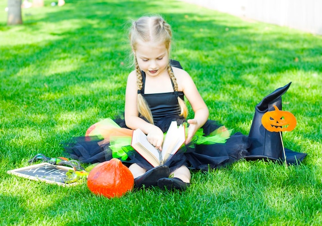 Uma menina vestida com uma fantasia de bruxa, um chapéu preto e batom preto nos lábios está sentada com um livro e segurando uma varinha mágica. dia das bruxas.