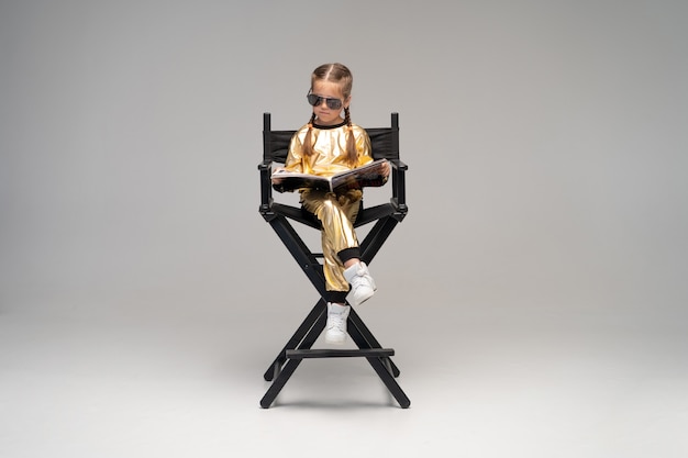 Uma menina vestida de ouro se senta em uma cadeira e lê um livro interessante. Isolado em uma superfície amarela