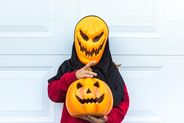 Uma menina vestindo uma máscara de abóbora assustadora de Halloween segurando uma abóbora sobre uma porta de garagem branca Conceito de fantasia de celebração carnaval terror medo e outono