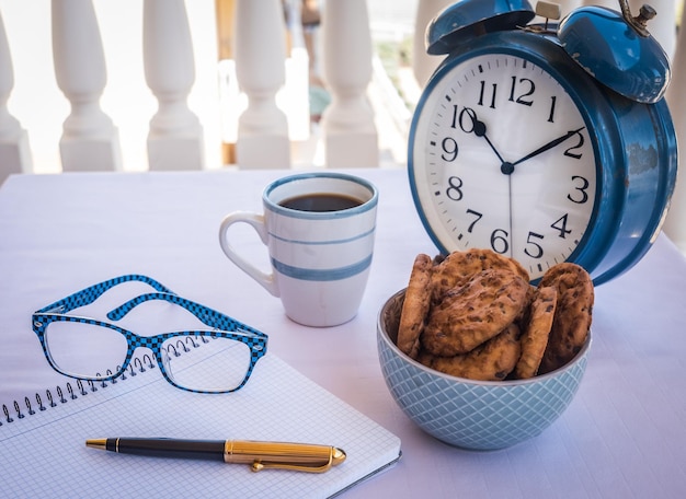 Uma mesa branca do lado de fora na varanda com uma xícara de café e alguns biscoitos com chocolate para uma pausa ou café da manhã Antigo despertador de metal azul