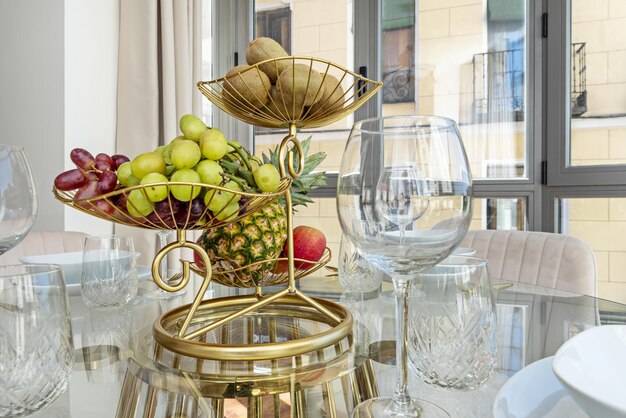 Uma mesa de jantar de vidro com uma fruteira dourada com uvas de abacaxi de vários tipos