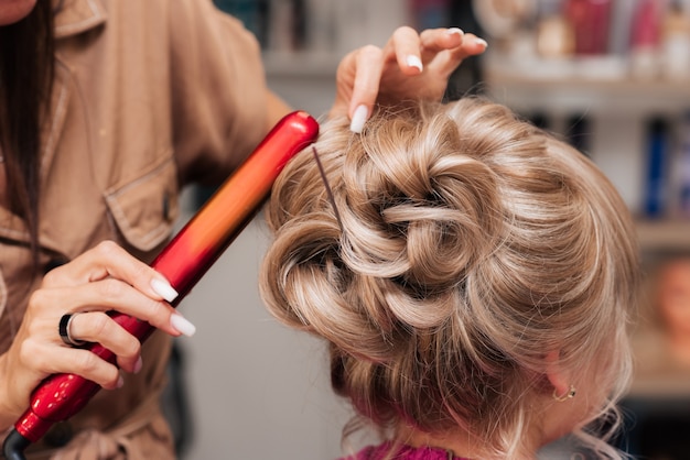 Uma moça cabeleireira faz o cabelo de uma cliente para uma festa com a ajuda de uma chapinha de ferro.