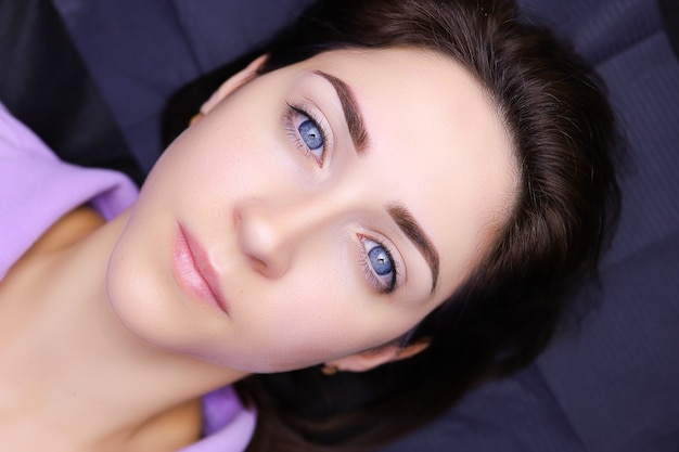 Uma modelo de olhos azuis deitada em um sofá de cosmetologia com as sobrancelhas tatuadas