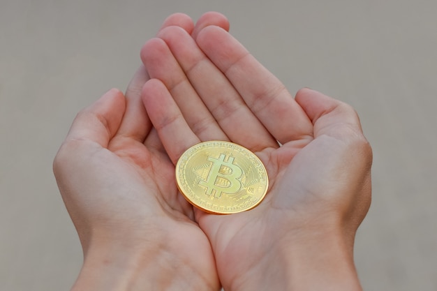 Uma moeda de bitcoin de criptomoeda dourada nas mãos da mulher alongada em uma superfície cinza. As mãos de uma mulher estão segurando uma grande moeda dourada de bitcoin.