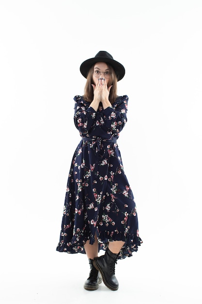 Uma morena com um longo vestido floral e um chapéu posa para a câmera no estúdio