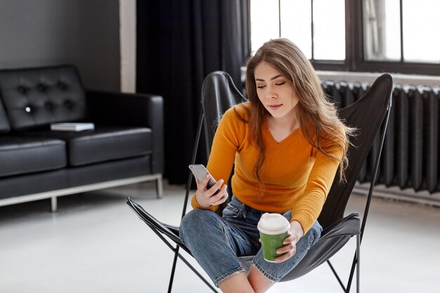 Uma morena linda com um suéter amarelo senta-se em uma elegante cadeira de couro preto, segura uma xícara de café e um telefone na mão. Trabalho, comunicação em casa, escritório