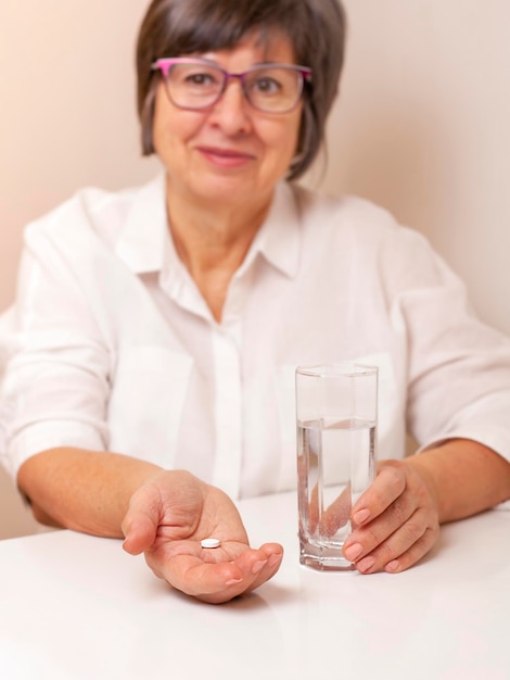 uma mulher adulta senta-se à mesa com um comprimido nas mãos e um copo de água. Concentre-se na pílula