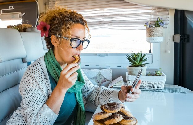 Uma mulher adulta sentada dentro de uma caravana desfruta de atividades de lazer indoor comendo donuts, bolos de açúcar e usando a conexão de telefone celular para navegar no mapa e escolher o próximo destino de viagem Vanlife Holiday