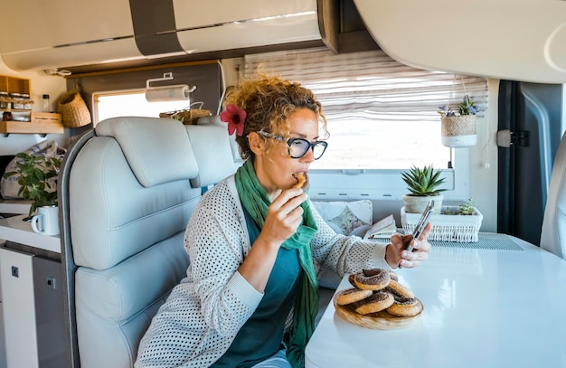 Uma mulher adulta sentada dentro de uma van de acampamento desfruta de atividades de lazer internas comendo donuts, bolos de açúcar e usando conexão de telefone celular para navegar no mapa e escolher o próximo destino de viagem Vanlife holiday