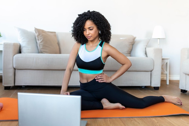 Uma mulher africana adulta faz exercícios de ioga e treinamento de força em um tapete em sua sala de estar Ela segue um vídeo de curso de exercícios on-line em seu laptop