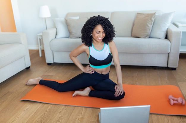 Uma mulher africana adulta faz exercícios de ioga e treinamento de força em um tapete em sua sala de estar Ela segue um vídeo de curso de exercícios on-line em seu laptop