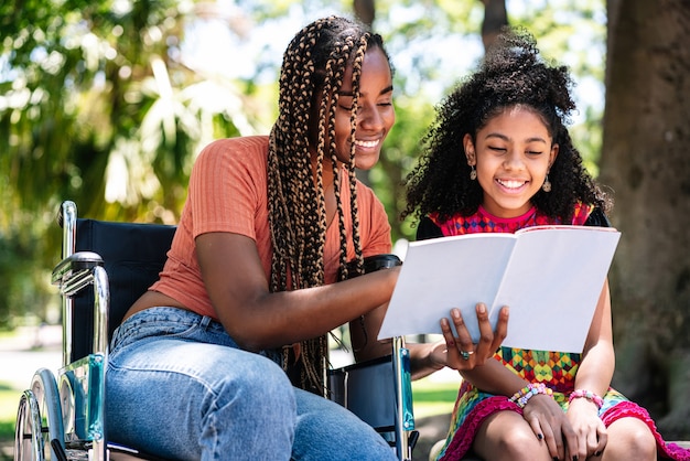 Uma mulher afro-americana em uma cadeira de rodas, curtindo um dia no parque com a filha enquanto lêem um livro