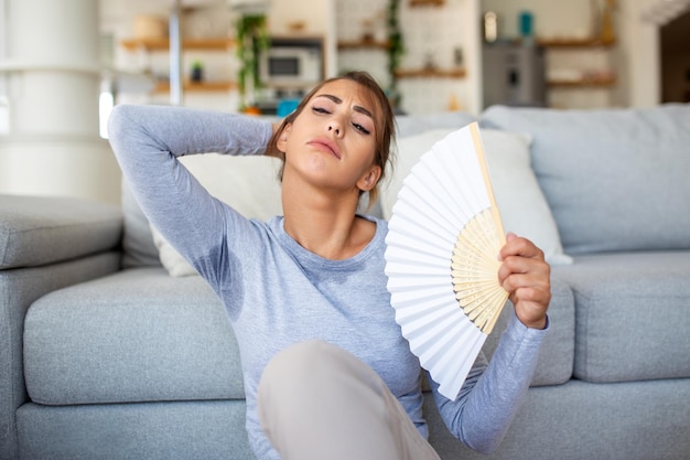 Uma mulher agitando um ventilador luta contra o superaquecimento e o calor do verão em casa Sua saúde e hormônios são afetados drenados pela exaustão devido à falta de ar condicionado O conceito de insolação