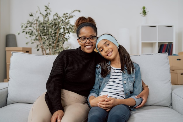 Uma mulher alegre e a filha sorridente abraçam-se sentadas no sofá no apartamento relaxando de embalar t