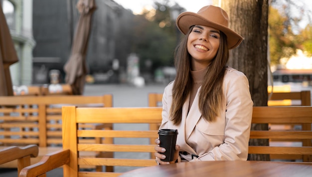 Uma mulher alegre na rua bebe café da manhã se senta em uma mesa. Copo descartável