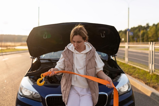 Uma mulher ao lado de um carro quebrado segura um cabo com um gancho para transportar um carro nas mãos